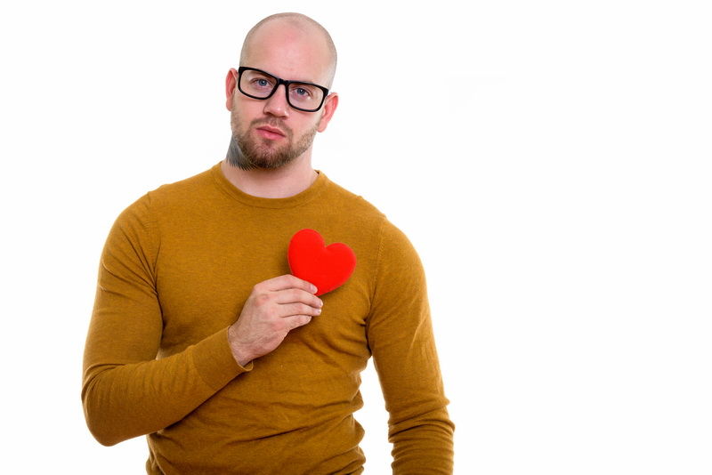 Studio shot of young bald muscular man holding red heart against chest ready for Valentine's day